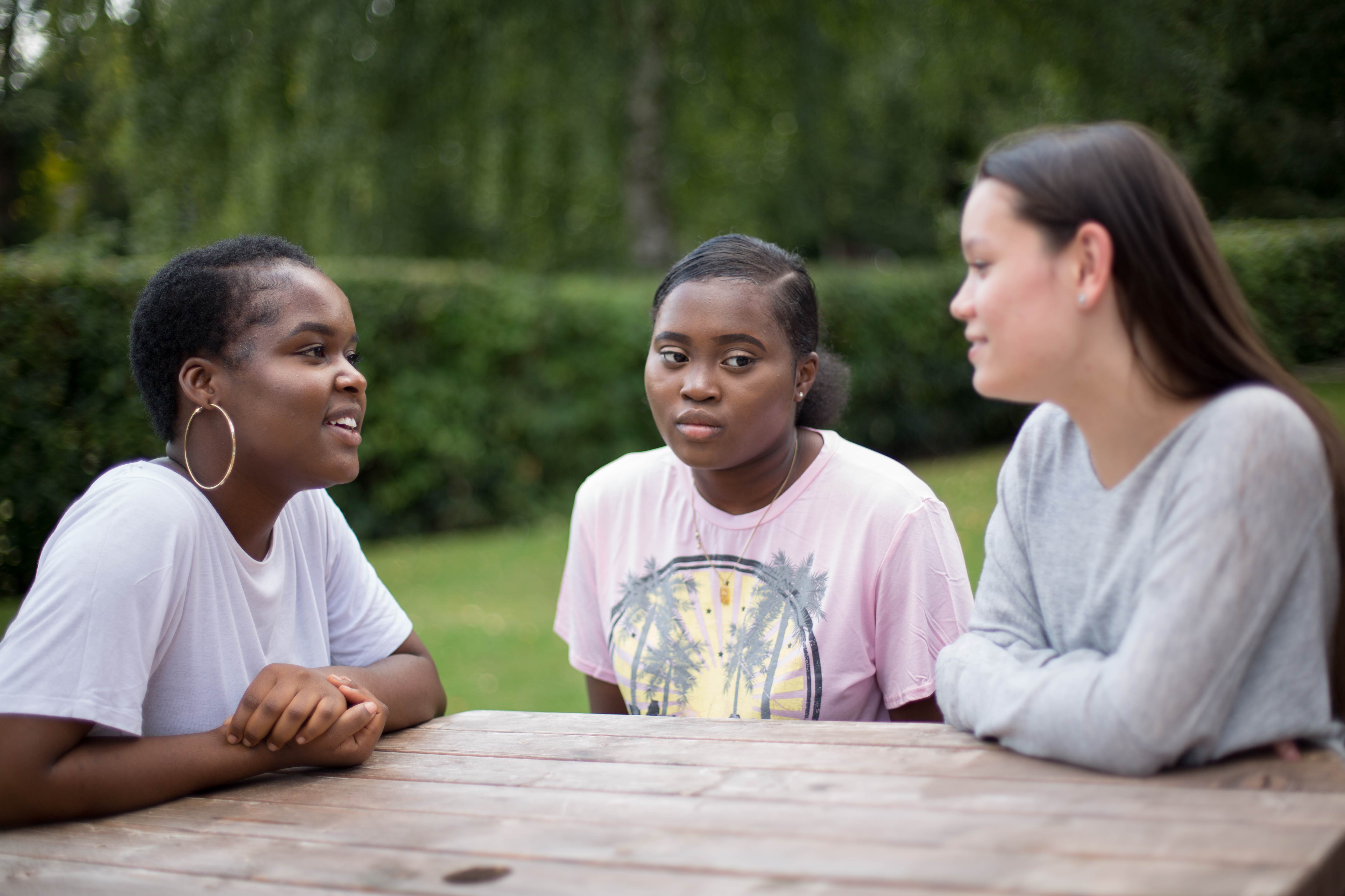 Three young people sitting at a table outdoors in a garden. They are having a conversation. Two are smiling, one has a neutral expression.