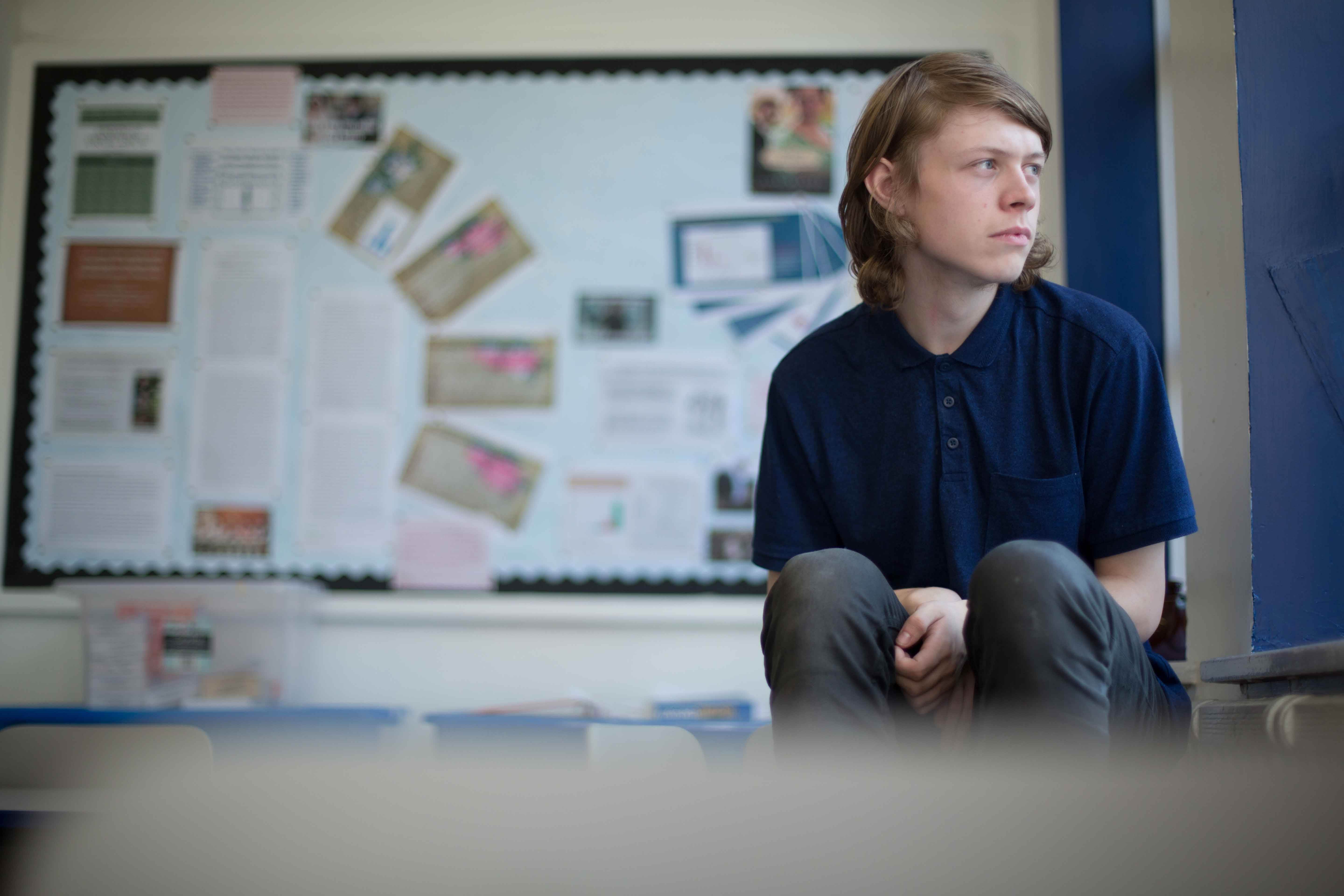 A teenage male with blonde hair in a blue t-shirt and grey jeans, sitting inside a secondary school classroom with a neutral expression. He is looking out of the window. 