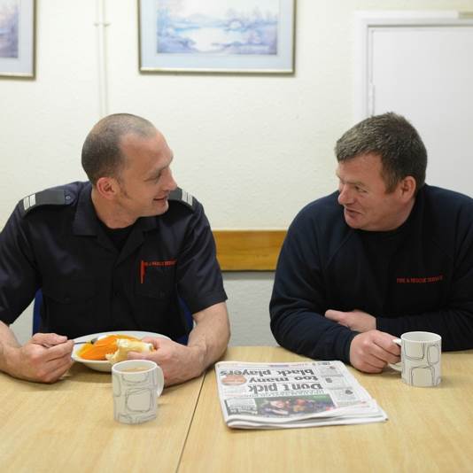 Two male firefighters chat at a table while one of them eats, a newspaper lies on the table between them and each has a mug