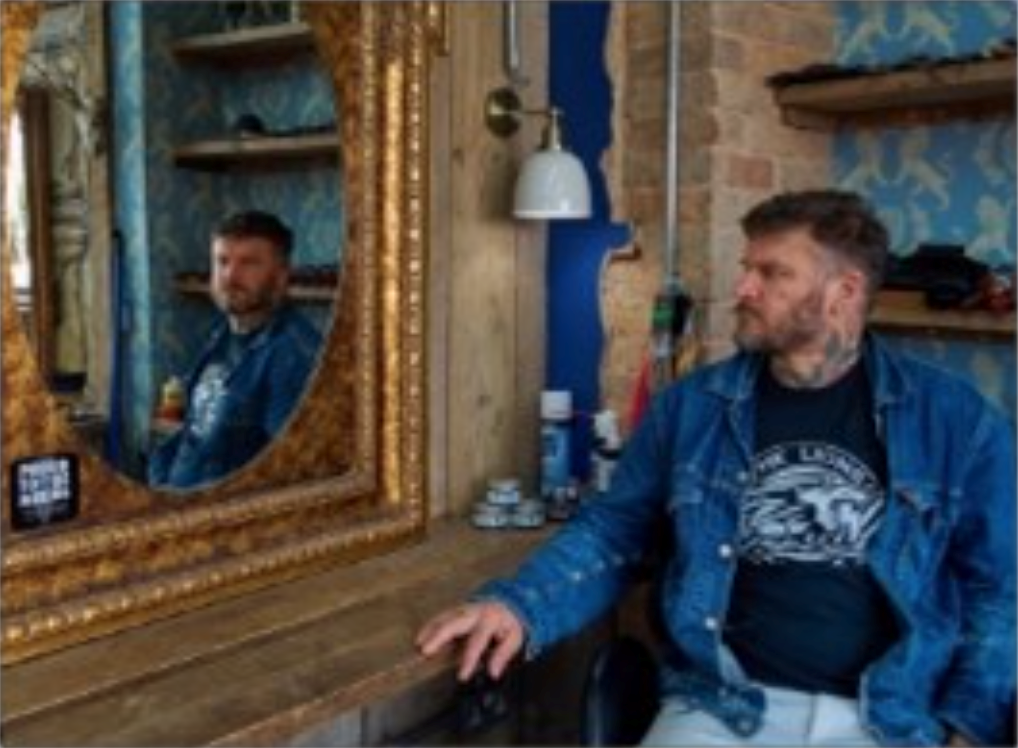 Male who is quite heavily tattooed sitting, looking at himself is a gold framed mirror
