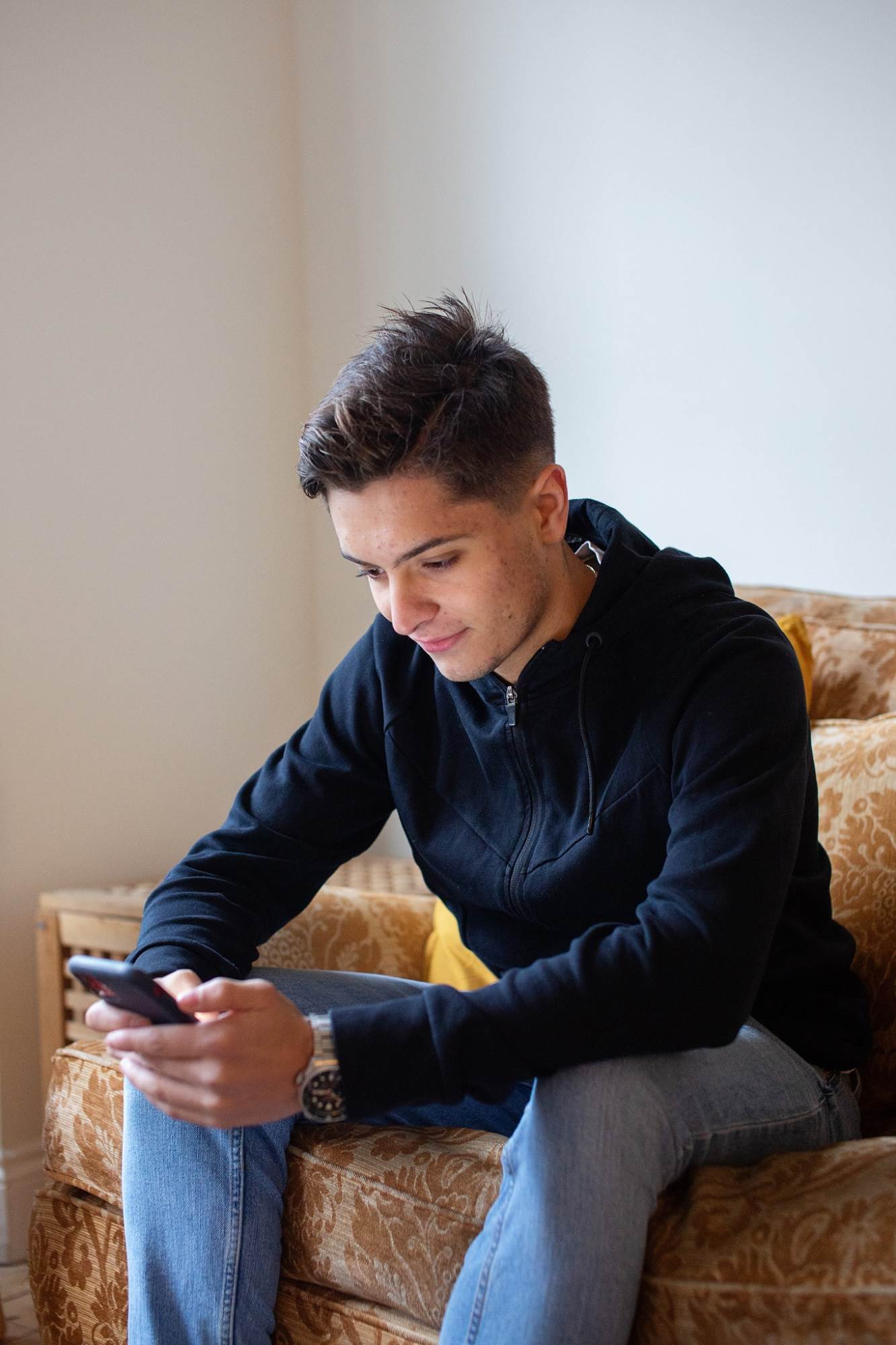 Teen Boy Smiling Looking At Mobile Content Sat On Sofa