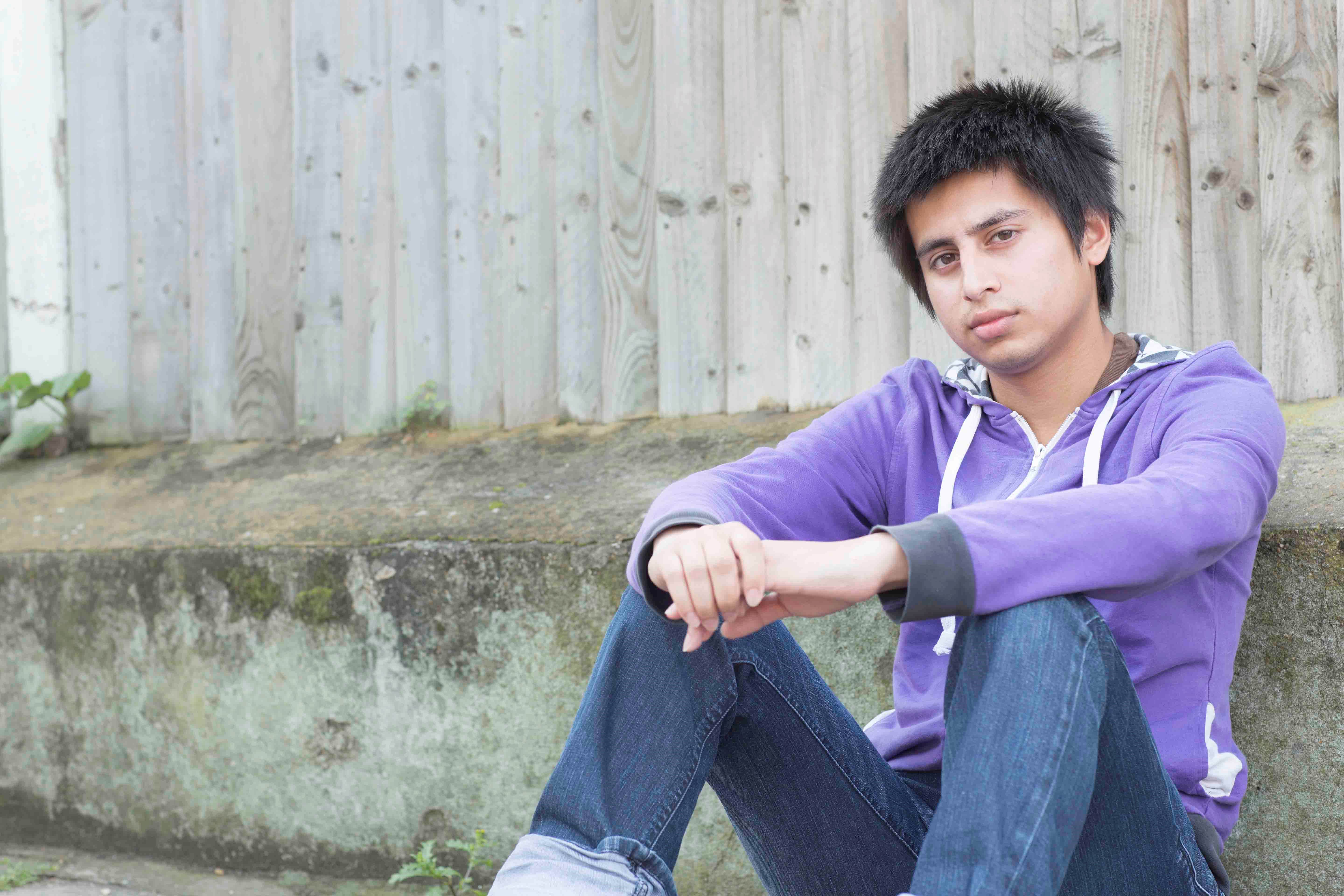 A young man in a purple hoodie and jeans with a neutral expression, sitting outside on the ground leaning against a fence.
