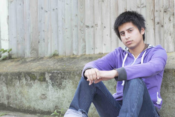 A young man in a purple hoodie and jeans with a neutral expression, sitting outside on the ground leaning against a fence. A young man in a purple hoodie and jeans with a neutral expression, sitting outside on the ground leaning against a fence.