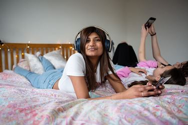 Teen Girl Wearing Headphones On Phone Lying On Bed Looks At Camera Smiling