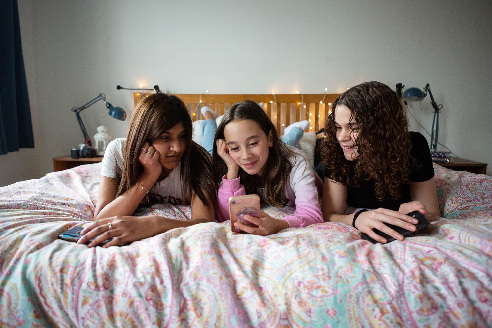 Two Teen Girls And Young Girl Smiling While Looking At Mobile Content On Bed