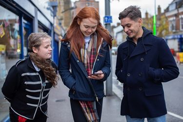 Two Teen Girls With Teen Boy Smiling At Mobile Content In Street