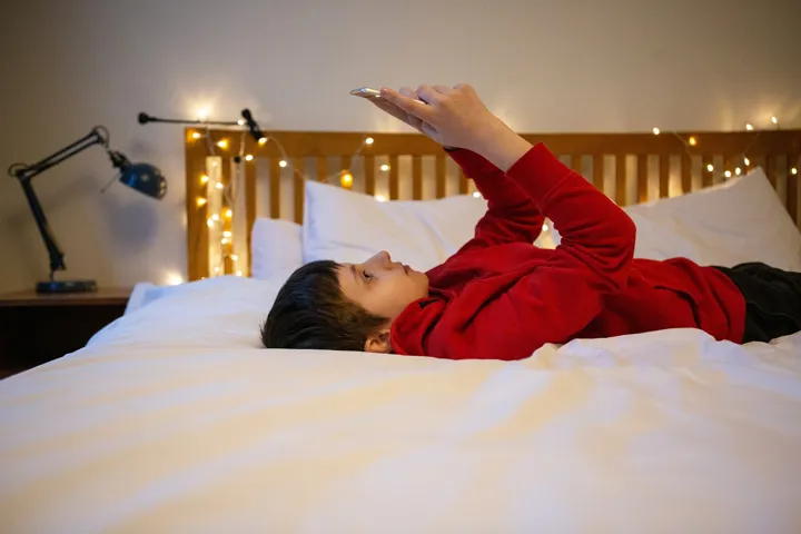 Young Boy Lying On Back On Bed Looking At Mobile Content Young Boy Lying On Back On Bed Looking At Mobile Content