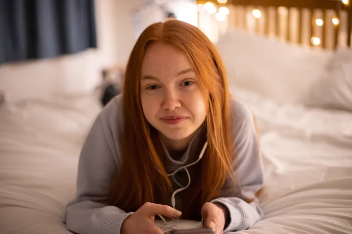 Young Girl Lies On Bed Using Phone And Smiling At Camera