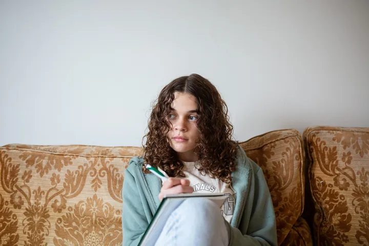 Young Girl Looks To The Left While Writing On Notebook Sat On Sofa Young Girl Looks To The Left While Writing On Notebook Sat On Sofa