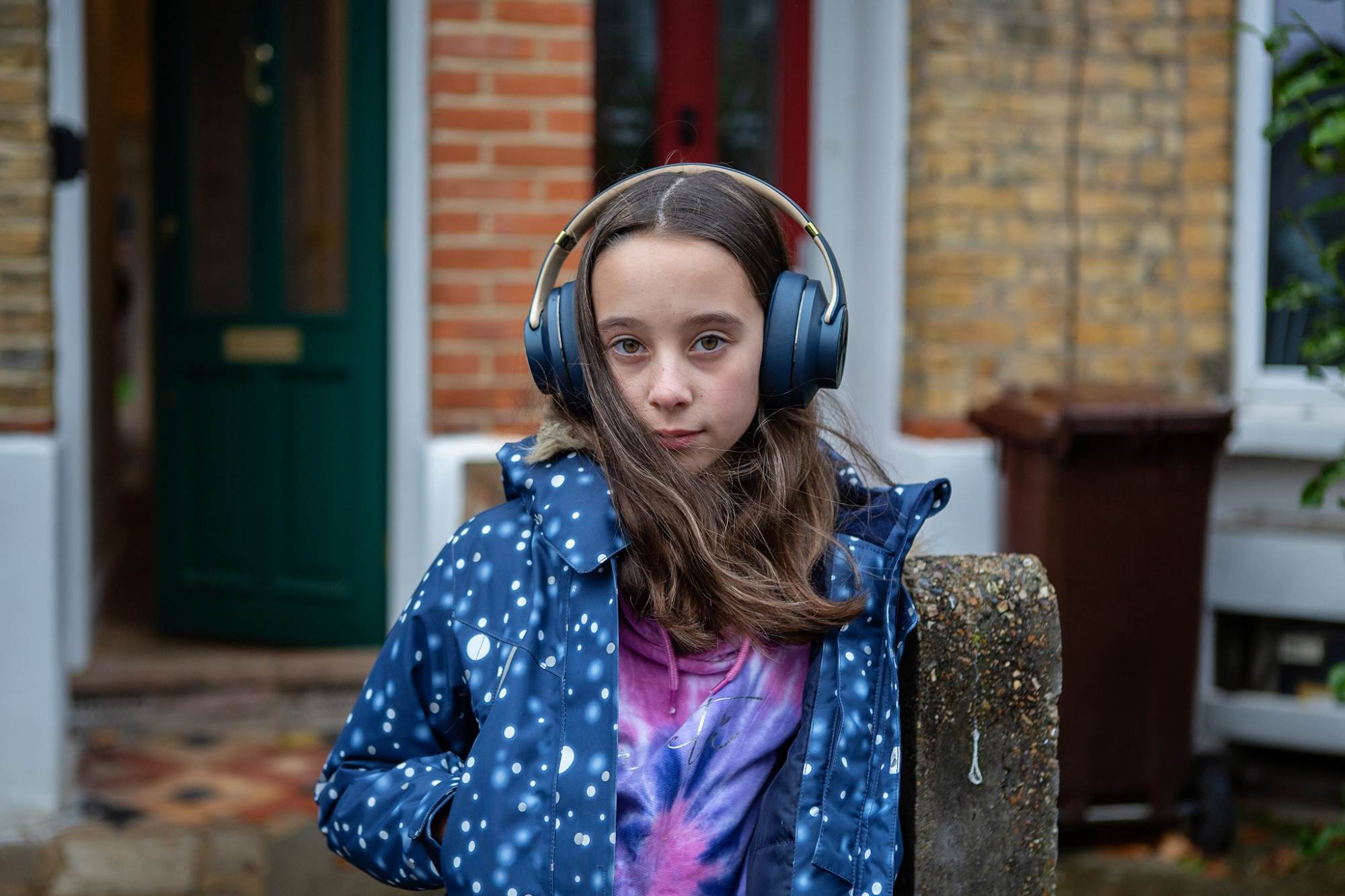 Young Girl Stands In Front Of House Listening To Headphones Looking At Camera