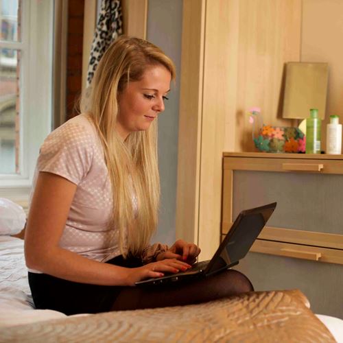 Woman sits on the side of her bed, looking at her laptop