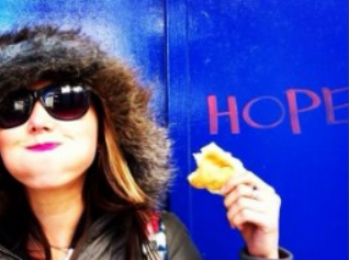 photo of woman wearing a fluffy hat and sunglasses, smiling and holding a bread roll, in front of a blue wall with the word "hope" on it