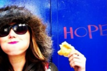 photo of woman wearing a fluffy hat and sunglasses, smiling and holding a bread roll, in front of a blue wall with the word "hope" on it