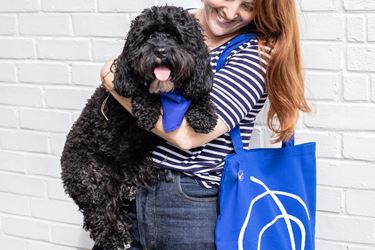 White woman with red hair and striped top, holding a cute fluffy black dog. The dog is wearing a blue Mind bandana, and the woman is carrying a blue Mind tote bag with a white squiggle. 