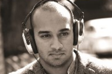 South Asian man with a shaved head and wearing headphones, with a neutral expression