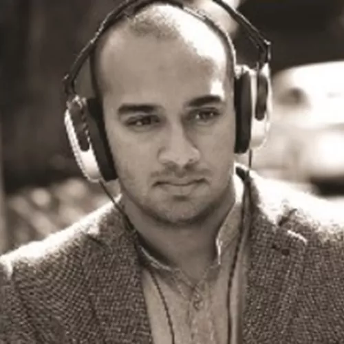 South Asian man with a shaved head and wearing headphones, with a neutral expression