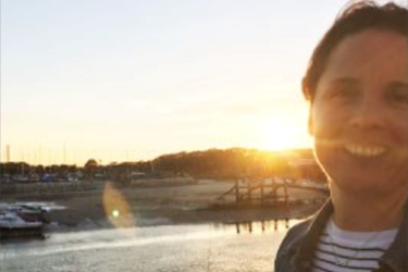 A woman looking at the camera with the sun setting behind her, with the sea, beach and docked boats.