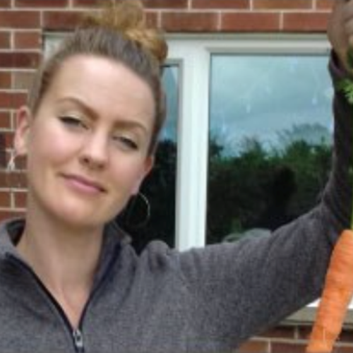 Woman holding a carrot up by the green leaves, and smiling at the camera