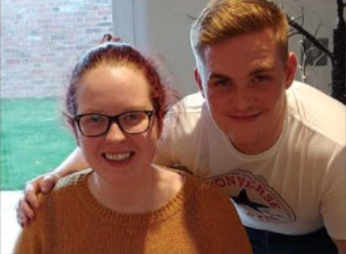 A young man crouching with his arm around a woman with pink, tied back hair. Both are smiling 