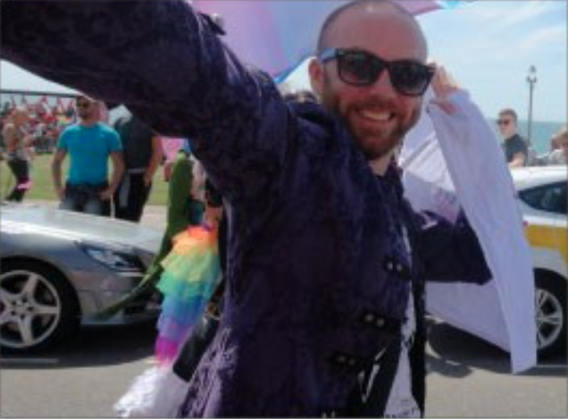 Male holding a flag with people and cars around him and an LGBT+ flag skirt
