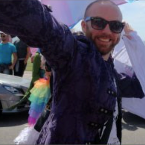 Male holding a flag with people and cars around him and an LGBT+ flag skirt