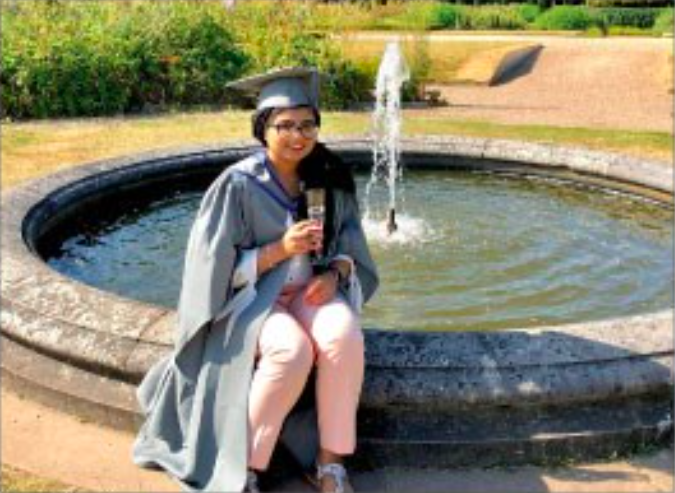 female wearing graduation robes and hat smiling sitting on the short wall of a water feature