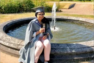 female wearing graduation robes and hat smiling sitting on the short wall of a water feature