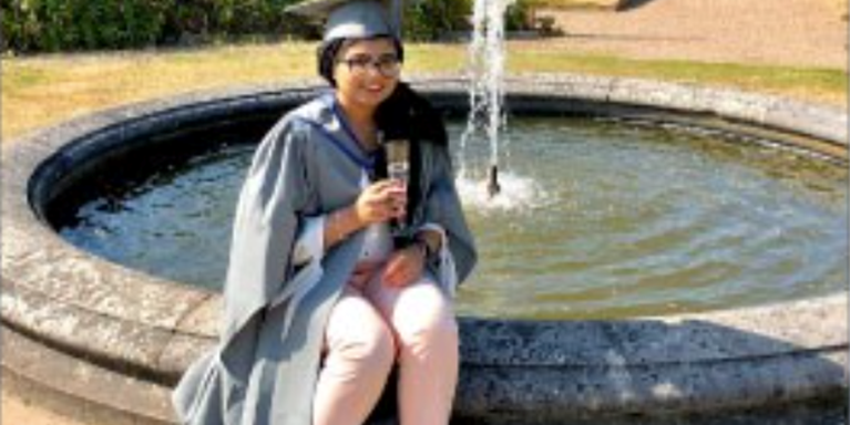 female wearing graduation robes and hat smiling sitting on the short wall of a water feature