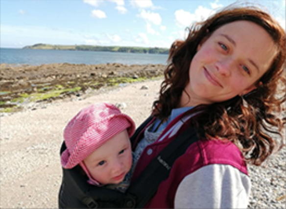 female smiling on a beach with a young child strapped to her front