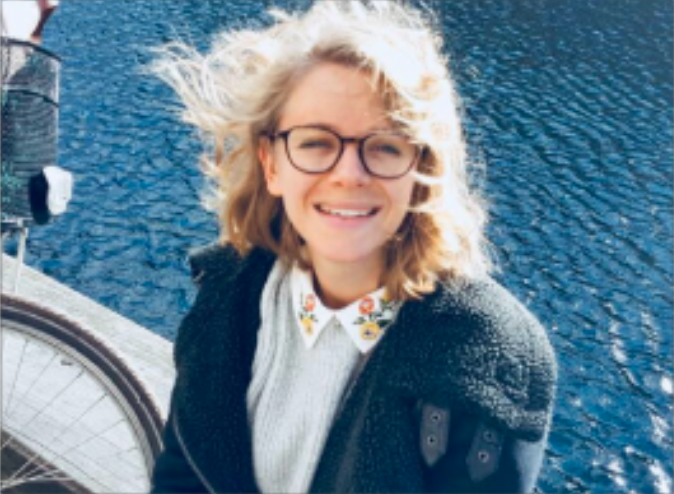 female smiling with her hair being flicked int he wind and is sitting in front of a body of water