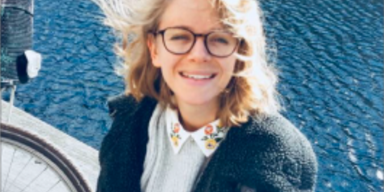 female smiling with her hair being flicked int he wind and is sitting in front of a body of water