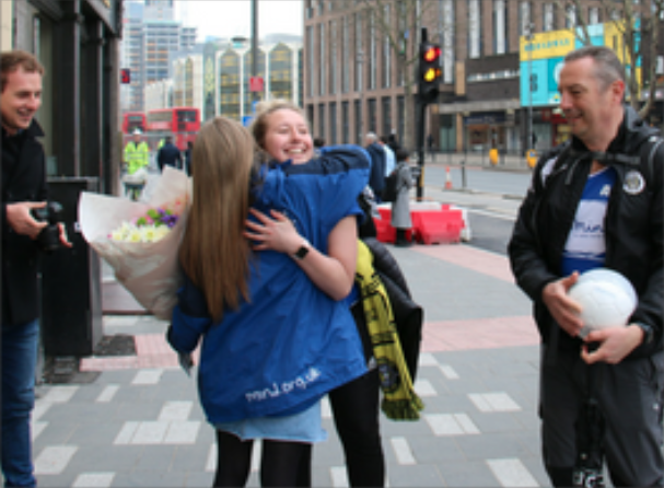 two females embracing on a pavement, one holding flowers 
