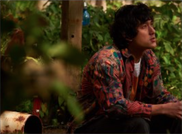 male leaning arms on knees and looking up, with green foliage around