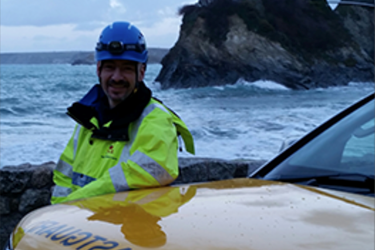 Male in high vis jacket standing by a car, with the sea behind him