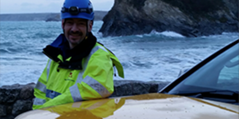 Male in high vis jacket standing by a car, with the sea behind him