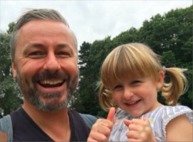 adult male holds a young female child close to him as she gives two thumbs up