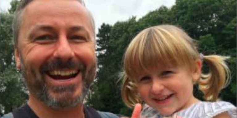 adult male holds a young female child close to him as she gives two thumbs up
