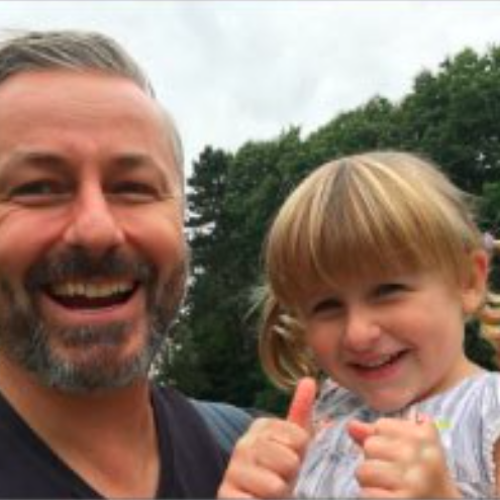 adult male holds a young female child close to him as she gives two thumbs up