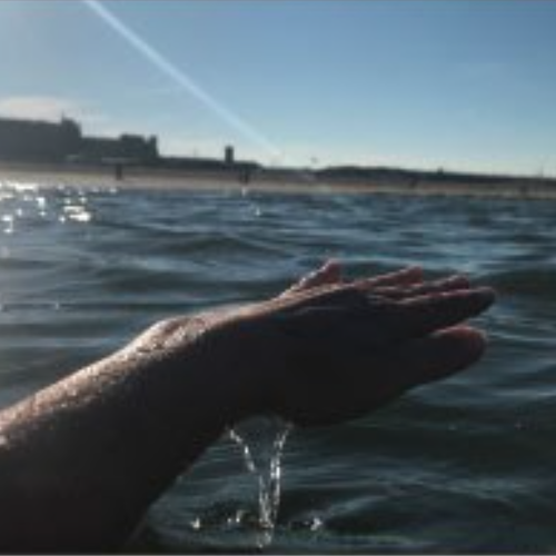 someone's hand lifted in the air, dripping with sea water, from the sea beneath it
