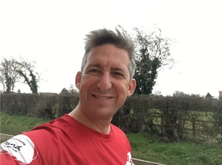 male smiling wearing a red top with trees behind it