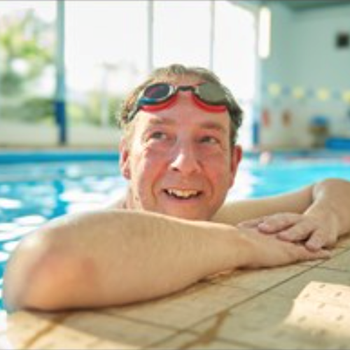 male wearing goggles leaning on the side of the swimming pool he is in