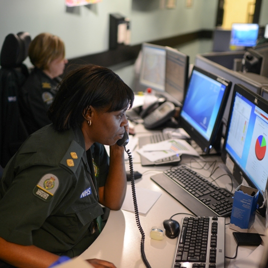A female Ambulance call handler on the phone in front of a bank of screens with her colleague in the background