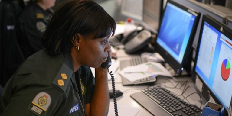 A female Ambulance call handler on the phone in front of a bank of screens with her colleague in the background