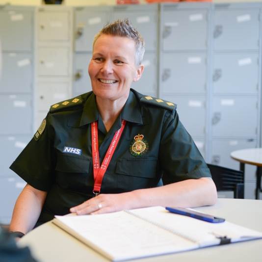 A smiling female Ambulance team member sits at a desk with an open book and pen in front of her and lockers in the background