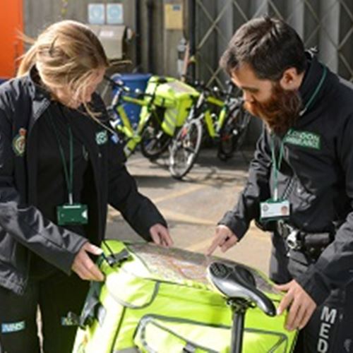 A male and a female Ambulance team member check a bright yellow box of equipment on the back of a bicycle
