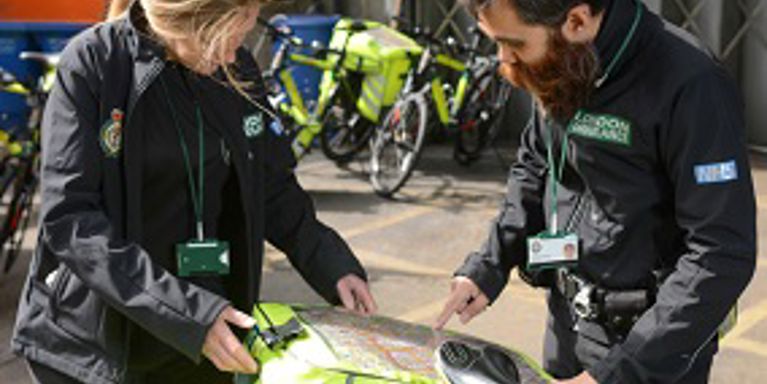 A male and a female Ambulance team member check a bright yellow box of equipment on the back of a bicycle
