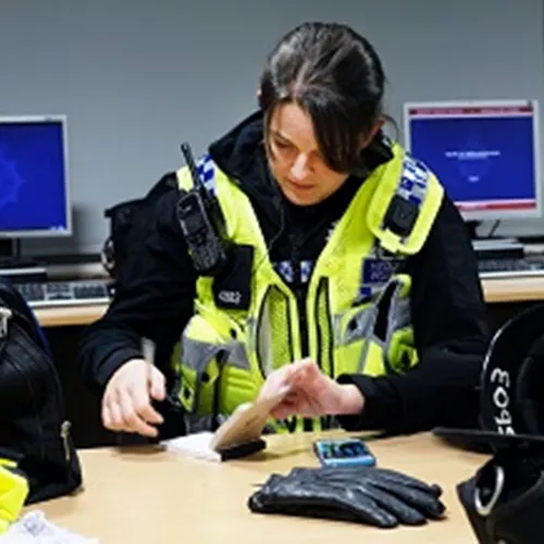 A female Police officer wearing a high viz vest and consulting her notebook at a table. Computer screens in the background.