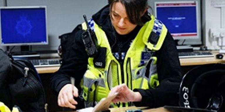 A female Police officer wearing a high viz vest and consulting her notebook at a table. Computer screens in the background.
