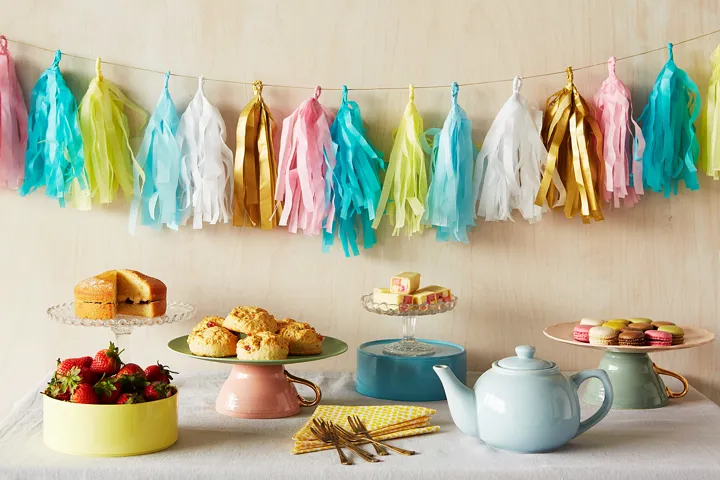 A kitchen table spread with baked treats, cakes, a bowl of fruit, napkins, cutlery, and a pot of tea. Behind the table, on the wall, there is a multi coloured paper garland.