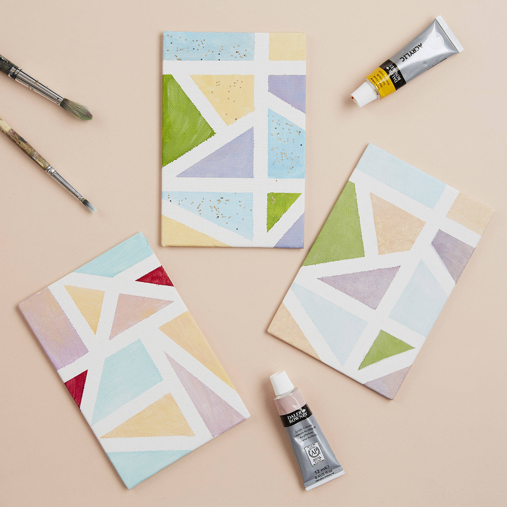 Three pieces of paper with geometric patterns painted onto them, with paint tubes and paintbrishes sitting beside them.